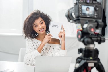 Neşeli Afro-Amerikan video blogcusunun seçici odak noktası diş telleri içinde dijital kameranın yanında etkili pazarlama harfleri olan bir defter tutması. 