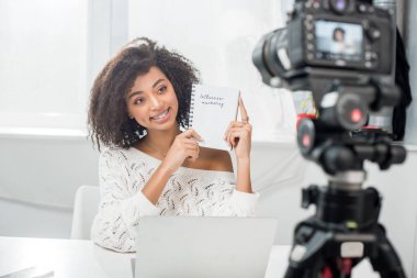 selective focus of cheerful african american video blogger in braces holding notebook with influencer marketing lettering near digital camera  clipart