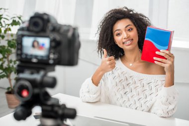 Dişlerinde renkli bir kutu tutan ve dijital kameranın yanında baş parmağını gösteren mutlu Afro-Amerikan etkisinin seçici odağı. 
