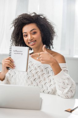 Diş teli takmış neşeli Afro-Amerikalı kız evde etkili pazarlama harfleri olan defterini işaret ediyor.