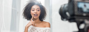 Panoramik çekim. Şaşırmış Afrikalı Amerikalı etkileyicinin diş telleriyle dijital kameranın yanındaki yüze dokunuşu. 