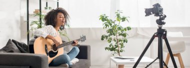 Panoramik çekimde, güzel Afro-Amerikan etkileyicisi diş telleriyle dijital kameranın yanında akustik gitar çalıyor. 