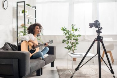 Diş teli takmış, dijital kameranın yanında akustik gitar çalan mutlu Afrikalı Amerikalı kız. 