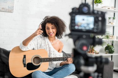selective focus of cheerful african american girl in braces holding acoustic guitar and pointing with finger at digital camera  clipart
