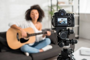 selective focus of digital camera with young african american girl in braces playing acoustic guitar and singing on display  clipart