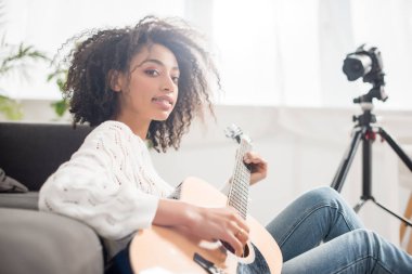 Genç ve kıvırcık Afrikalı diş telli Amerikalı kız dijital kameranın yanında akustik gitar tutuyor. 
