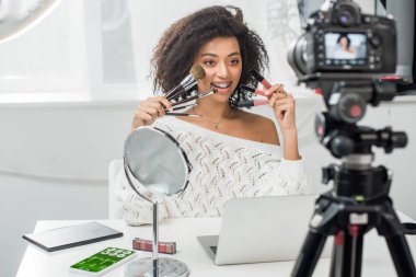 Diş teli takmış, dudak parlatıcısı ve spor telefonu yanında spor takip cihazı ve dijital kamerayla kozmetik fırçalar tutan mutlu Afro-Amerikan etkileyicinin seçici odak noktası.