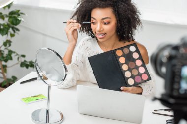 En iyi alışveriş uygulaması ve dijital kamera ile akıllı telefonun yanında göz farı süren neşeli Afro-Amerikan video blogcusunun seçici odağı 