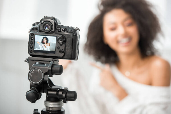 selective focus of digital camera with happy african american influencer in braces pointing with finger at credit card on display 