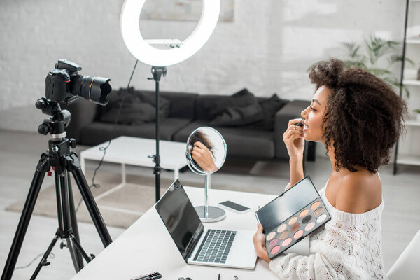 side view of happy african american influencer in braces holding eye shadow palette near digital camera 
