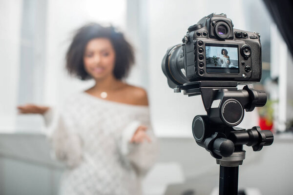 selective focus of digital camera with attractive african american influencer gesturing while talking on display 