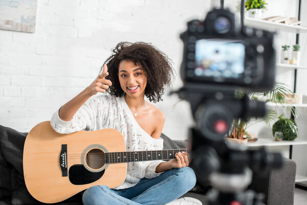 selective focus of cheerful african american girl in braces holding acoustic guitar and pointing with finger at digital camera 