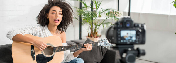 панорамный снимок молодой африканской девушки в брекетах, играющей на акустической гитаре и поющей возле цифровой камеры
 