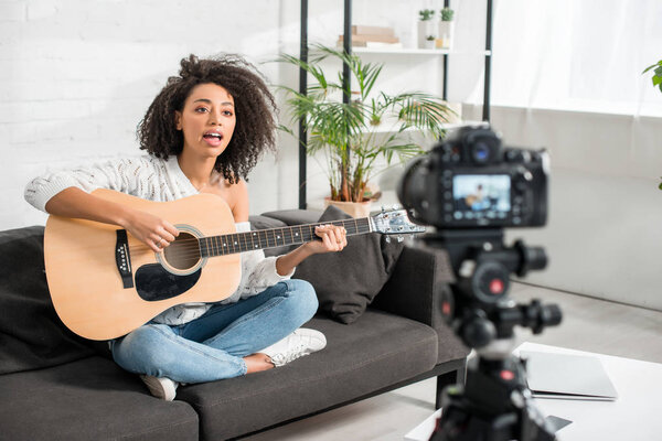 селективный фокус молодой африканской девушки в брекетах, играющей на акустической гитаре и поющей возле цифровой камеры
 