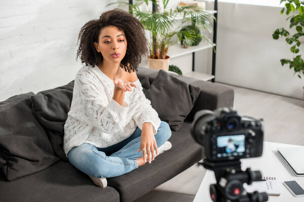 selective focus of young african american influencer sitting on sofa and sending air kiss near digital camera 