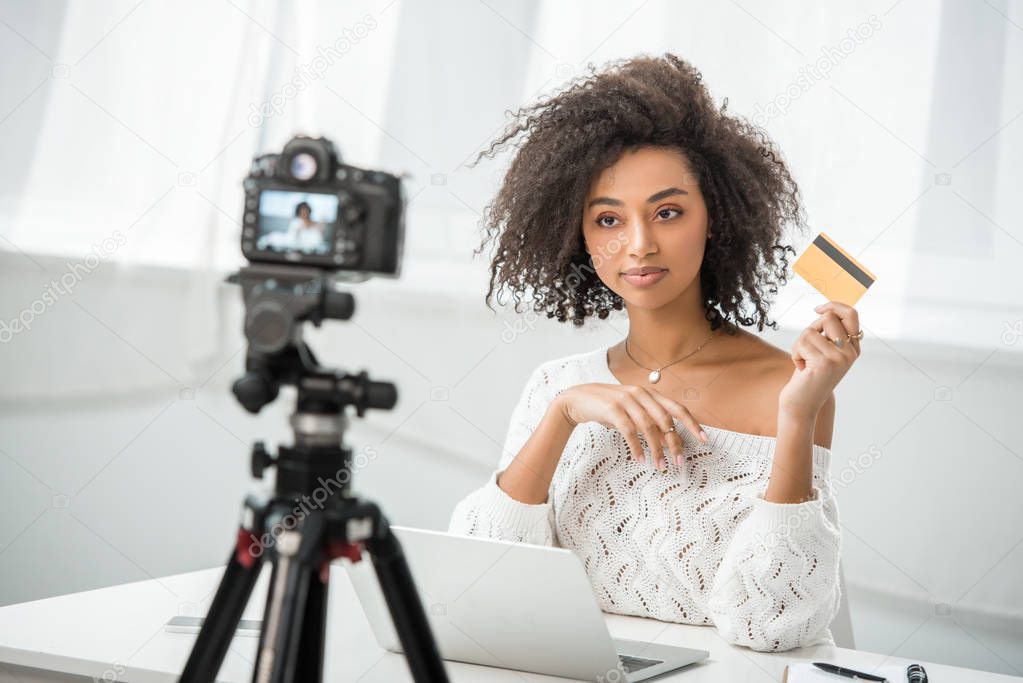 selective focus of african american influencer holding credit card near laptop and looking at digital camera 
