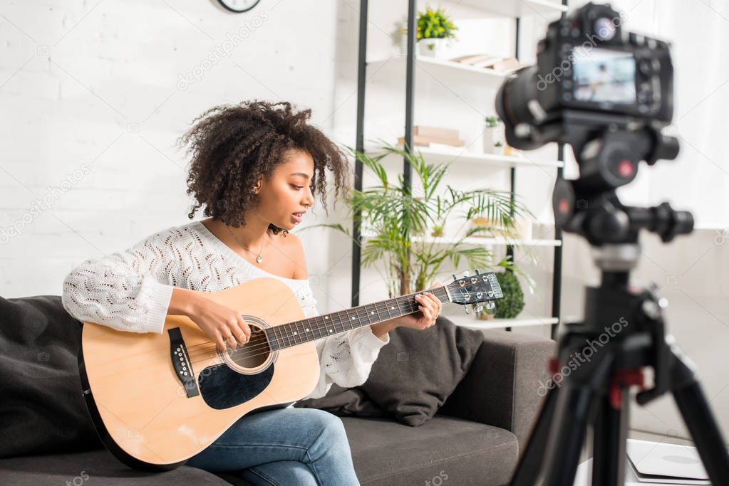selective focus of curly african american girl playing acoustic guitar near digital camera 