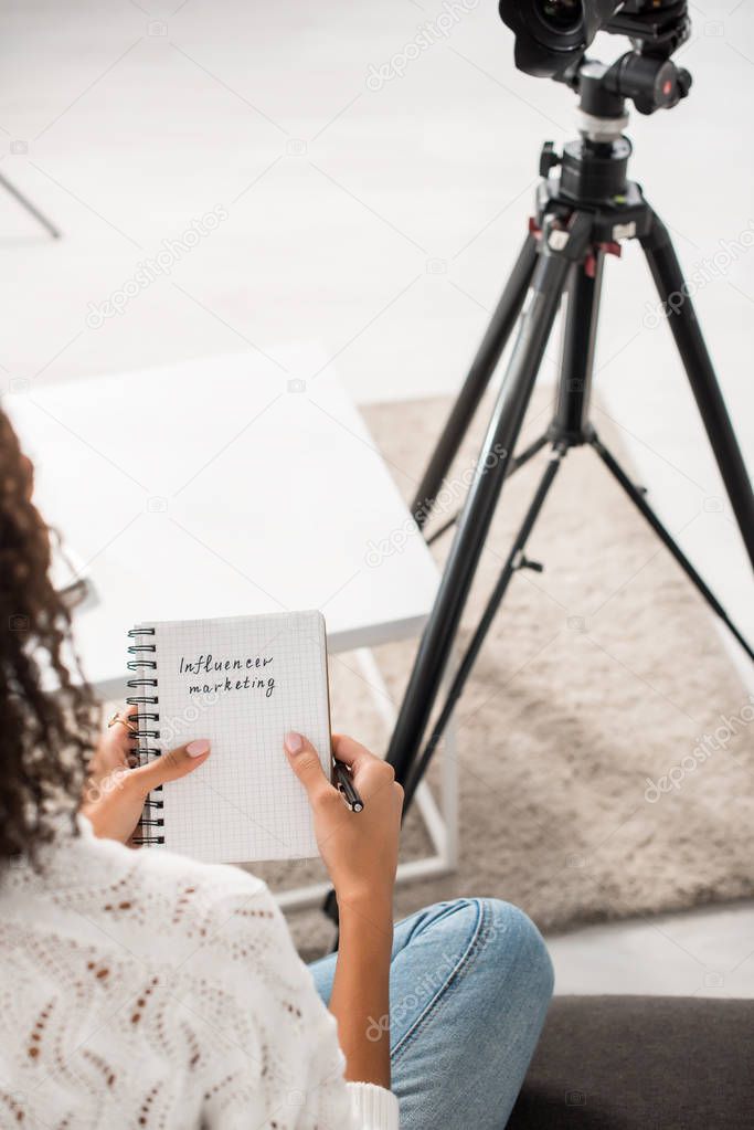 cropped view of curly african american girl holding notebook with influencer marketing lettering at home 