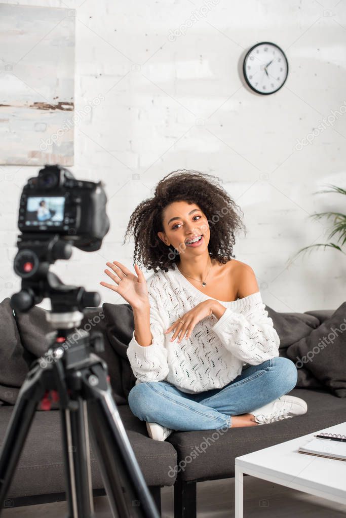 selective focus of happy african american influencer in braces waving hand and talking near digital camera 