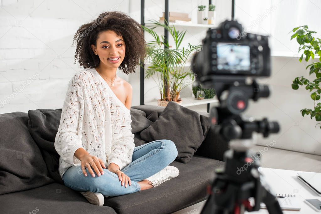 Selective focus of happy african american influencer sitting on sofa and looking at digital camera