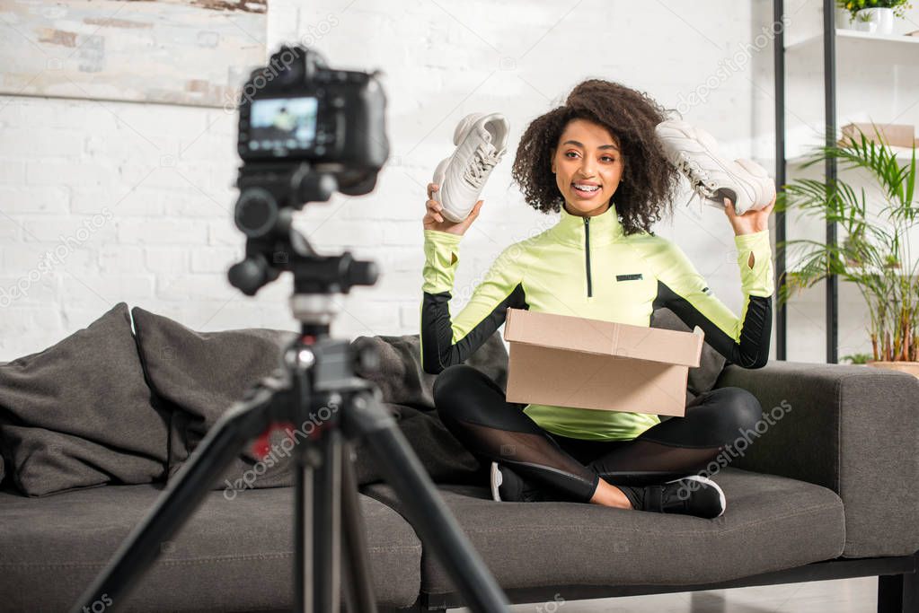 selective focus of happy and sportive african american influencer in braces holding new sneakers near box and digital camera 