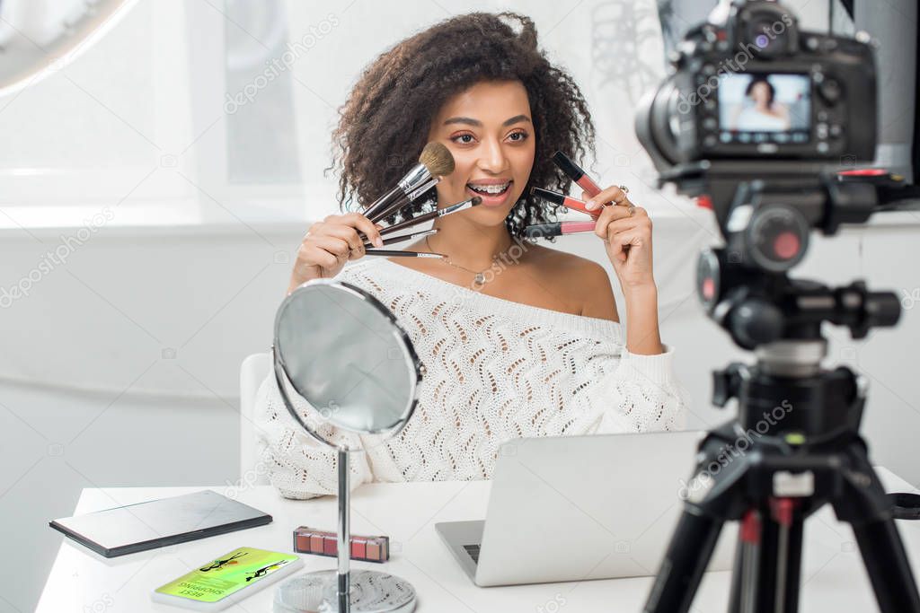 selective focus of happy african american influencer in braces holding lip glosses and cosmetic brushes near smartphone with best shopping app and digital camera