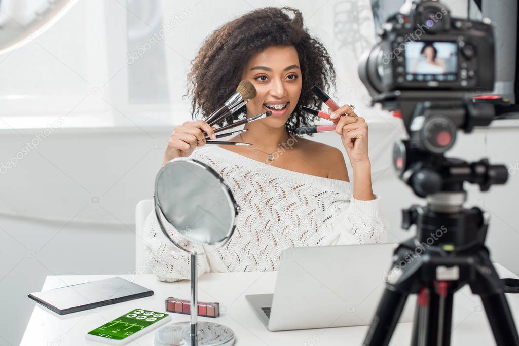 selective focus of happy african american influencer in braces holding lip glosses and cosmetic brushes near smartphone with fitness tracker and digital camera