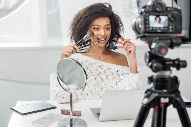 Akıllı telefonun yanında dudak parlatıcısı ve kozmetik fırça tutan ve ekranda online harita ve dijital kamera olan mutlu Afro-Amerikan etkisinin seçici odak noktası.