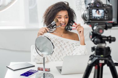 Diş teli takmış, dudak parlatıcısı ve akıllı telefonun yanında tıbbi takip cihazı ve dijital kamerayla kozmetik fırçalar tutan mutlu Afro-Amerikan etkileyicinin seçici odak noktası.