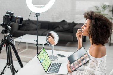 Mutlu Afro-Amerikalı etkileyicinin yan görüntüsü göz farı paletini ekranda harita ve dijital kamerayla birlikte dizüstü bilgisayarın yanında tutuyor 