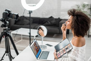 KYIV, UKRAINE - DECEMBER 10, 2019: side view of african american influencer holding eye shadow palette near laptop with couchsurfing website and digital camera  clipart