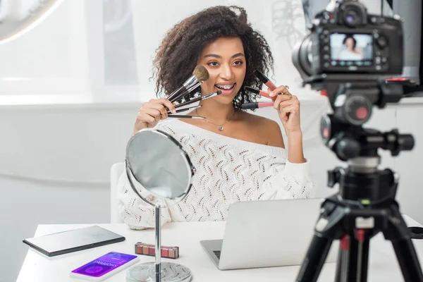 快乐的非洲裔美国影响者在智能手机附近拿着唇彩和化妆品刷 用购物软件和数码相机选择性地聚焦在焦点上 — 图库照片