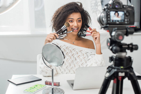 selective focus of happy african american influencer in braces holding lip glosses and cosmetic brushes near smartphone with map on screen and digital camera