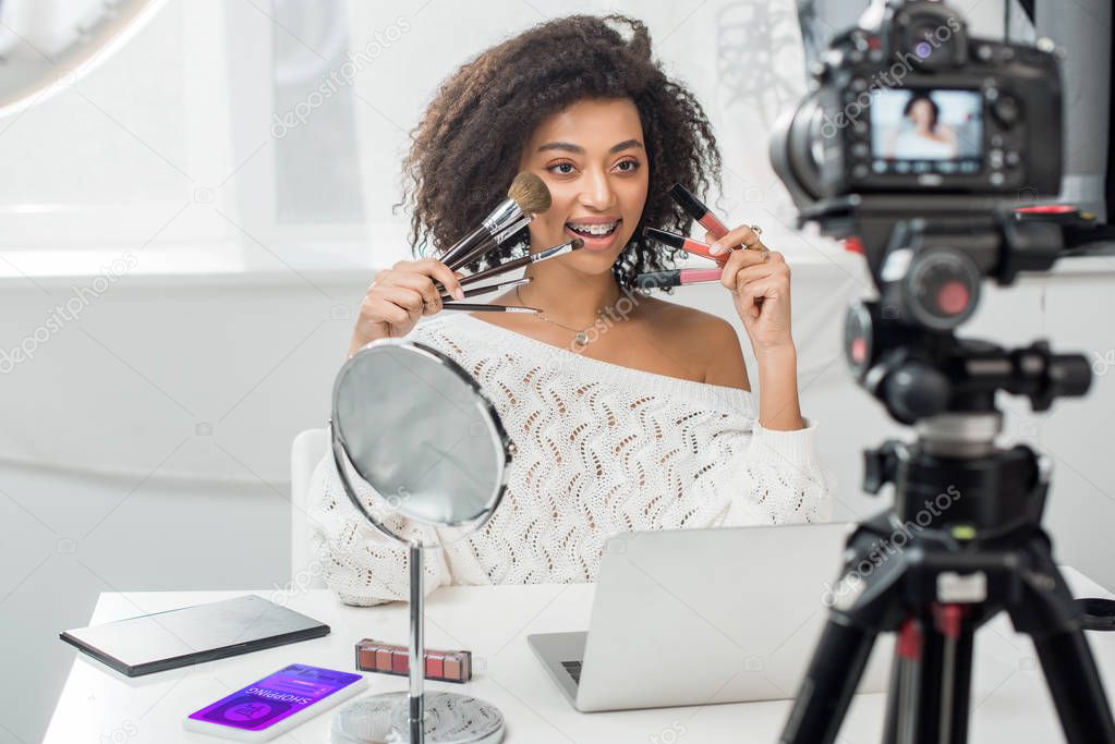 selective focus of happy african american influencer in braces holding lip glosses and cosmetic brushes near smartphone with shopping app and digital camera