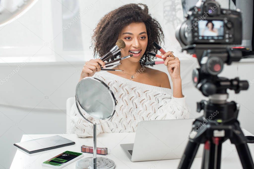selective focus of happy african american influencer in braces holding lip glosses and cosmetic brushes near smartphone with booking app and digital camera