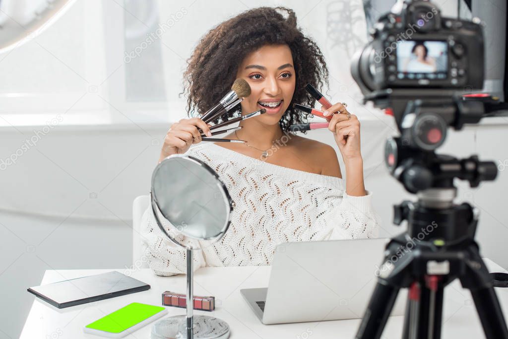 selective focus of happy african american influencer in braces holding lip glosses and cosmetic brushes near smartphone with green screen and digital camera