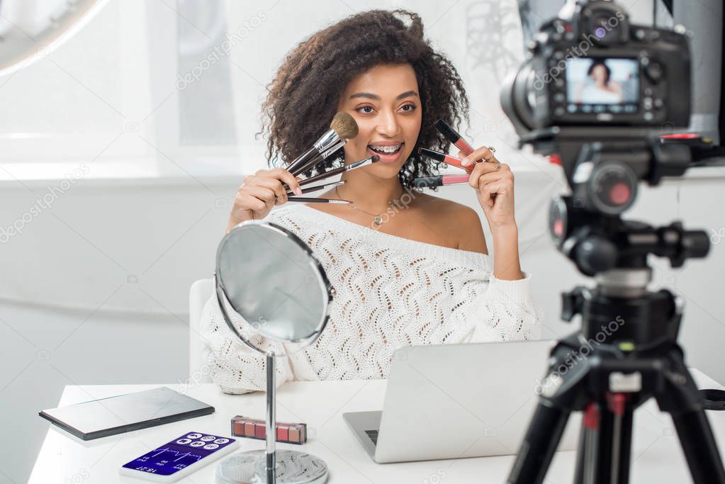 selective focus of happy african american influencer in braces holding lip glosses and cosmetic brushes near smartphone with medical tracker and digital camera