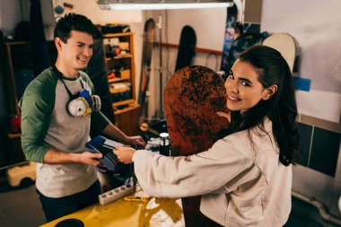 Terminalde çalışan ve gülümseyen bir kadın kredi kartıyla ödeme yapıyor ve tamir atölyesinde snowboard tutuyor. 