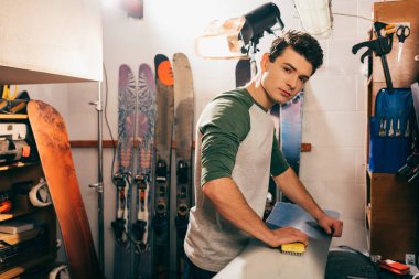 İşçi snowboard 'da fırça kullanıyor ve tamirhanedeki kameraya bakıyor. 