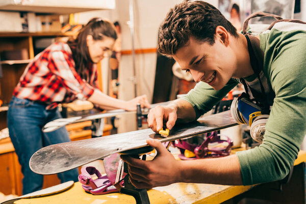selective focus of smiling worker using box cutter on ski in repair shop