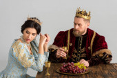 rozrušená královna a král s korunami sedí u stolu izolované na šedé