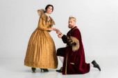king with crown bending on knee and holding hand of smiling queen on grey background 