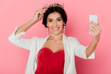 Taçlı gülümseyen kadın pembe arka planda akıllı telefon tutuyor. 