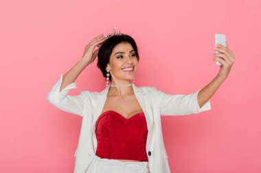 Taçlı gülen kadın pembe arka planda selfie çekiyor. 