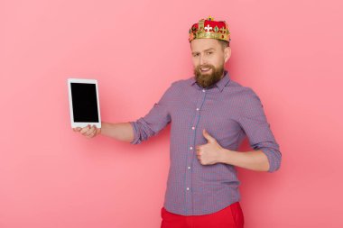 Taçlı gülümseyen adam pembe arka planda dijital tablet taşıyor. 