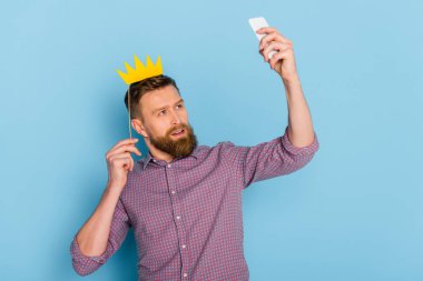 man holding paper crown and taking selfie on blue background  clipart