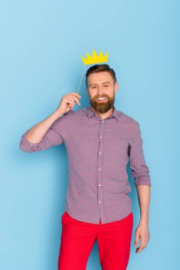 smiling and handsome man holding paper crown on blue background  clipart