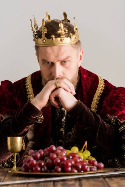 thoughtful king with crown sitting at table isolated on grey