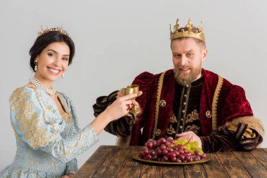 smiling queen and king with crowns clinking with cups isolated on grey