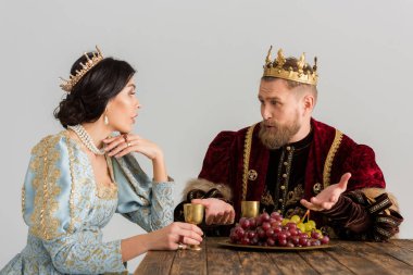 queen and king with crowns sitting at table and talking isolated on grey